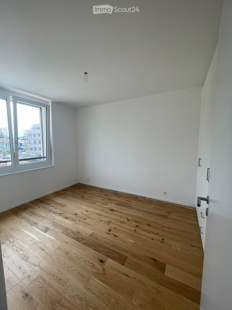 Empty room with light wooden flooring and white walls, featuring a large window with a view of the city and a door with silver handles.