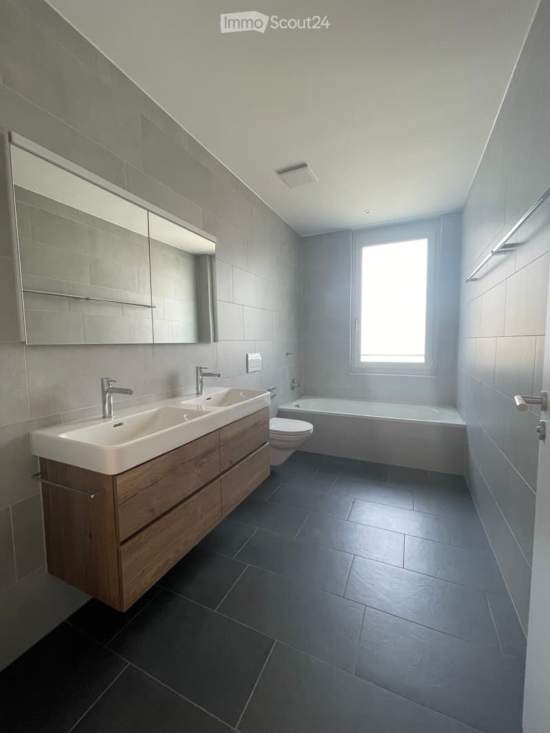 Modern bathroom with double sink, wooden vanity, large mirror, toilet, bathtub, and window providing natural light.