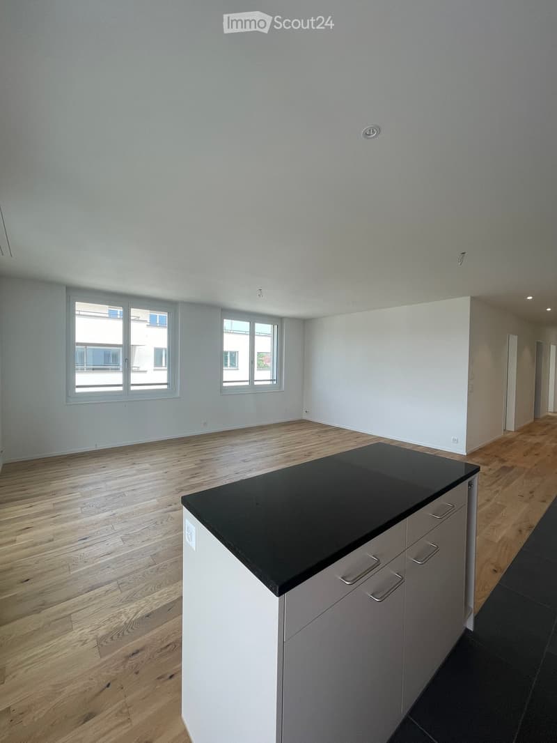 A modern, unfurnished room with light wooden flooring, white walls, and large windows. A kitchen island with a black countertop and white cabinets is visible.
