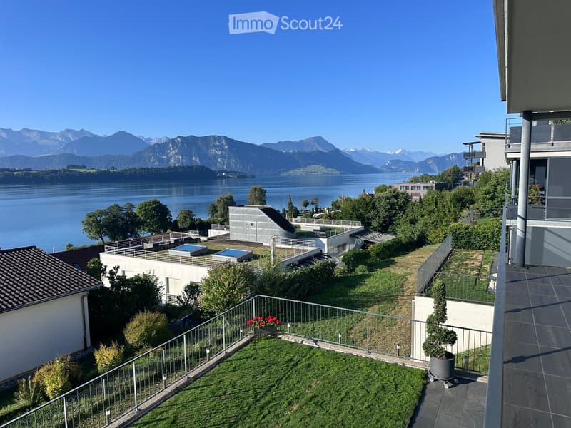 Garden area with a view of a lake and mountains, featuring a small lawn, potted plants, and a modern building with a balcony.