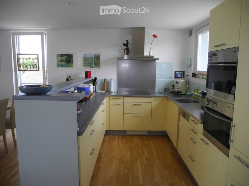 Modern kitchen with light yellow cabinets, grey countertops, and stainless steel appliances including an oven, fridge, and sink. Features a central island with a bowl and vase, and a window with a view of greenery.