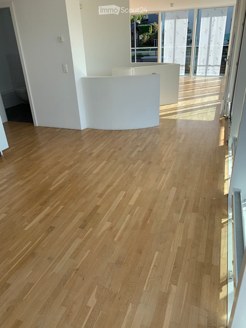 Empty room with light wooden flooring, white walls, and a curved white partition. Large windows and glass doors lead to an outdoor area.