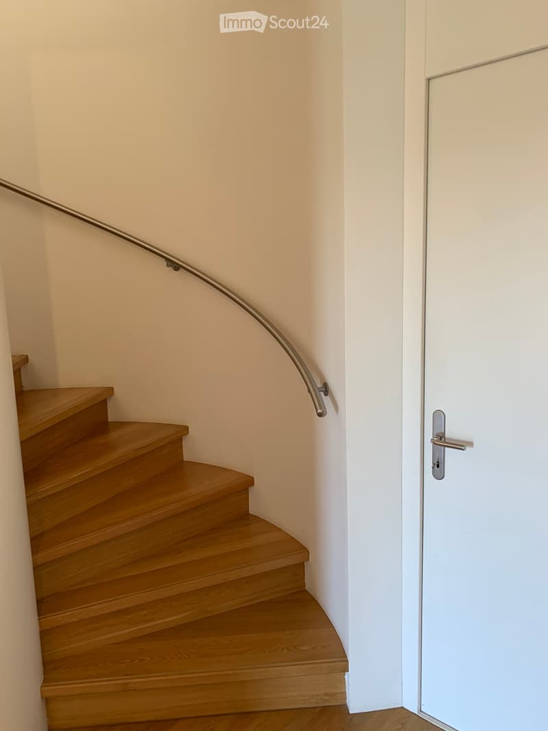 Wooden staircase with a curved metal handrail leading up, adjacent to a white door with a silver handle.