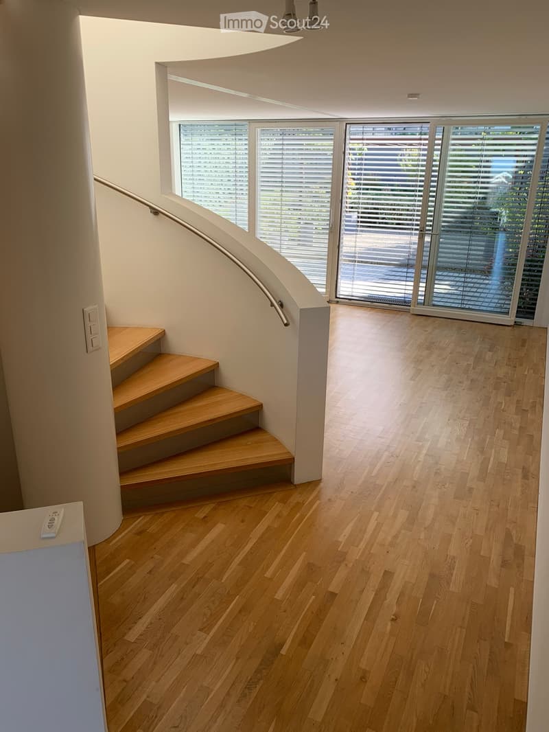 Modern hallway with wooden flooring, a wooden staircase with a metal handrail, and large windows with blinds leading to an outdoor area.