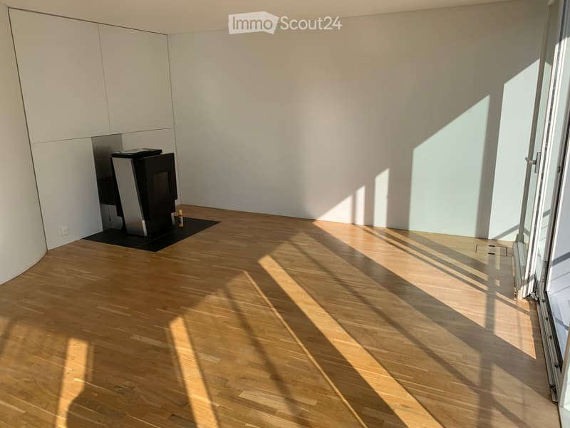 Empty room with light wooden flooring, white walls, and large windows allowing natural light to enter. A small black fireplace is visible in the corner.