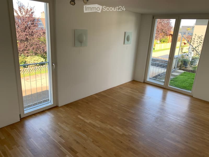 Empty room with wooden flooring, large sliding glass doors leading to a balcony and garden, and two wall-mounted decorative items.