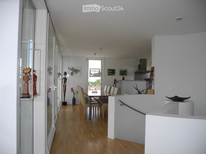 Open-plan dining and kitchen area with wooden flooring, modern white walls, and large windows. Features a dining table with chairs, a kitchen counter, and decorative items including a statue and framed artwork.