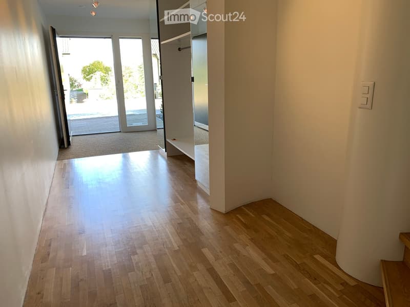 Bright hallway with wooden flooring, white walls, and a staircase leading upwards. A doorway leads to a carpeted area with large glass doors opening to an outdoor space.