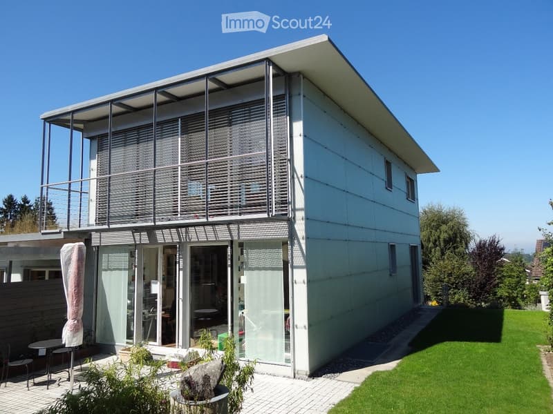 Modern two-story house with white exterior, large windows, and a balcony on the upper level. The ground floor features a covered patio area with outdoor furniture and plants.