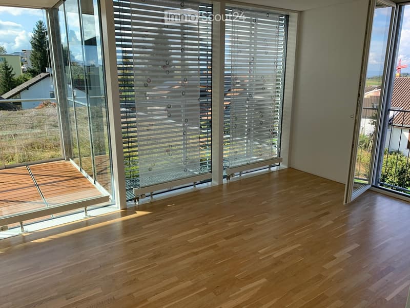 Empty room with wooden flooring and large windows with blinds, overlooking a balcony area.