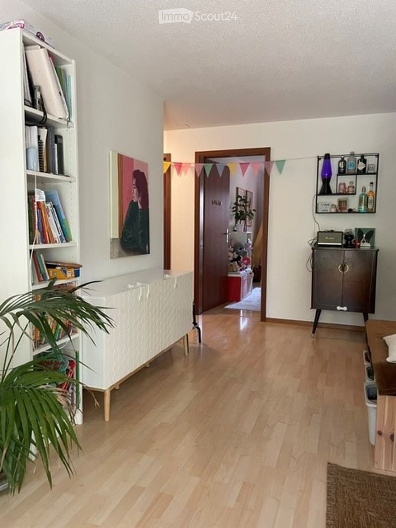 Bright, open living space with light wooden flooring, white walls, and a mix of modern and cozy furnishings including a bookshelf, a painting, a small table, and a potted plant.