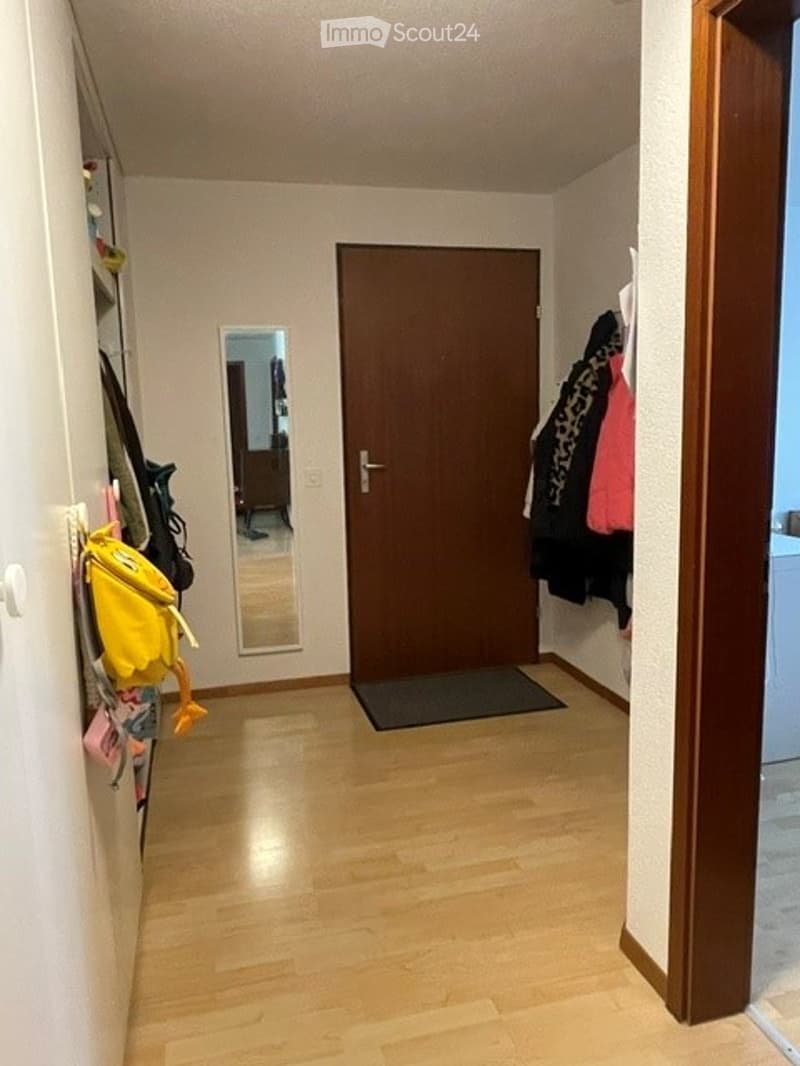 A narrow hallway with wooden flooring, a closed wooden door, a small mirror, and a coat rack with jackets and bags.