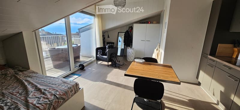 Bright, modern room with a bed, sofa, small table, and chair. Large sliding glass door opens to a balcony with outdoor seating. Built-in closet and kitchen area visible.