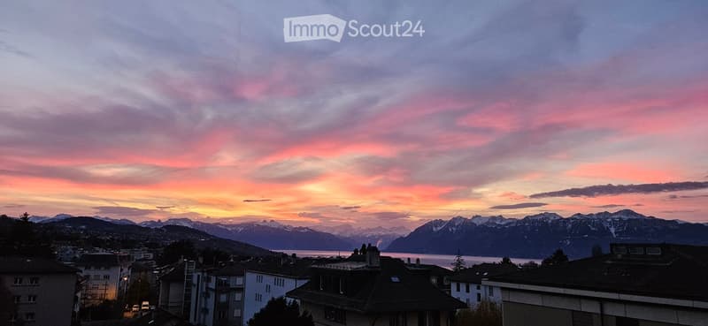 View of a cityscape at sunset with mountains and a lake in the background.