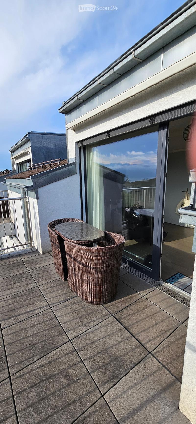 Balcony with tiled floor, glass door leading inside, two wicker chairs and a small table.