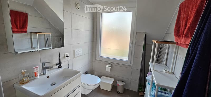 Modern bathroom with a white sink, chrome faucet, toilet, mirror, and shelves holding towels and toiletries. The room features grey tiled walls and a window providing natural light.