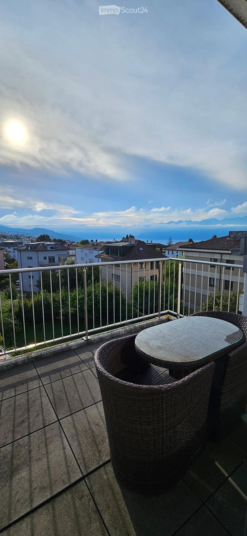 Balcony with a round glass table and two wicker chairs, overlooking a scenic view of a city and mountains.