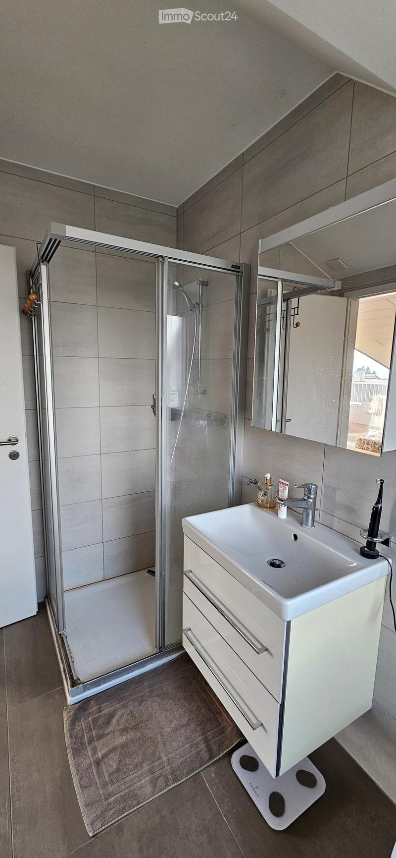 Modern bathroom with a white sink, mirror, and glass-enclosed shower. Includes a towel on the floor and a small bottle on the sink.
