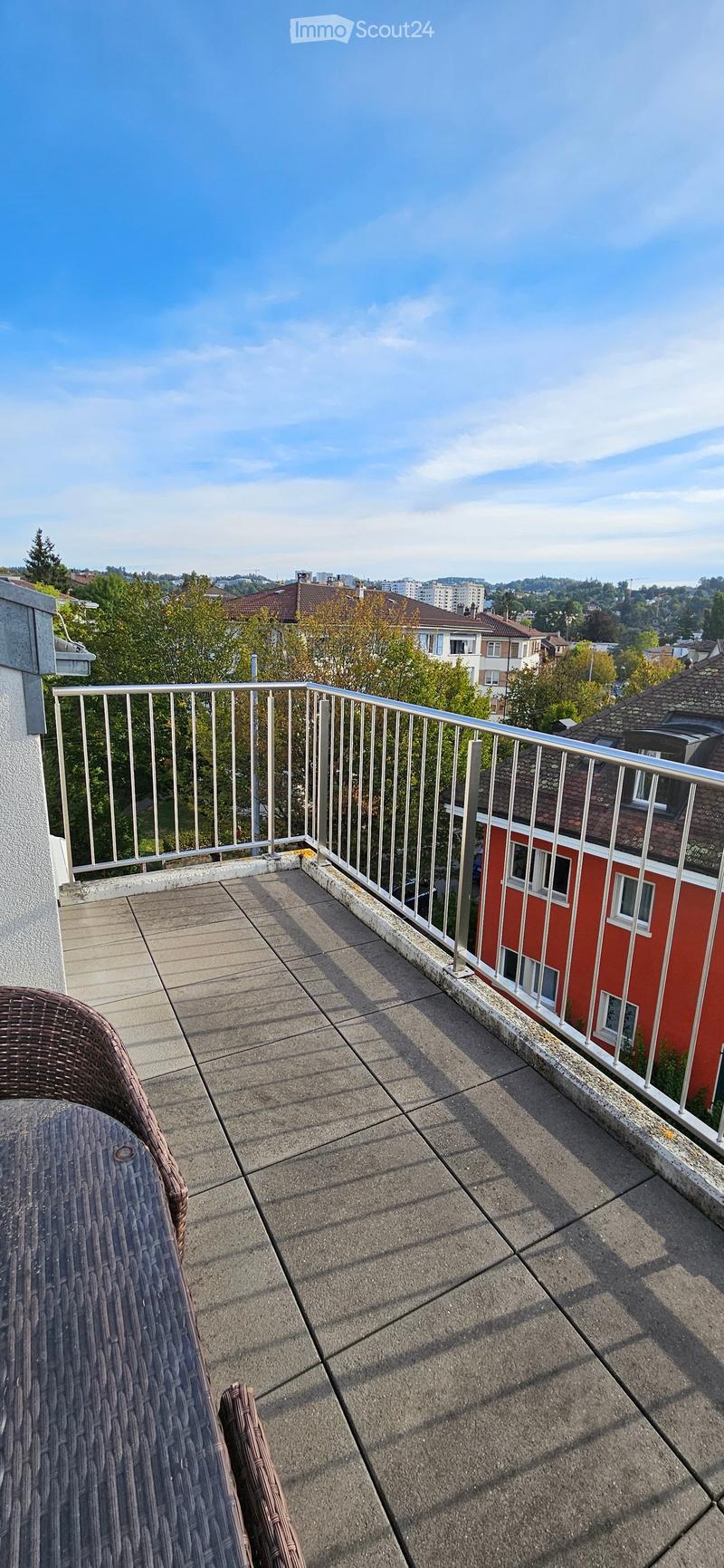 Balcony with a concrete floor, metal railing, and a view of the city and trees. Includes a wicker table and chair.