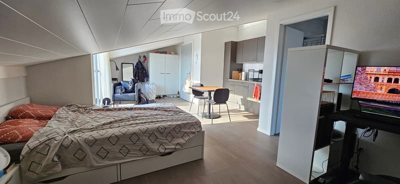 A compact, well-lit room with a bed, a small kitchen area, a chair, a table, and a laptop on a desk. The room features modern furniture and a cozy, organized layout.