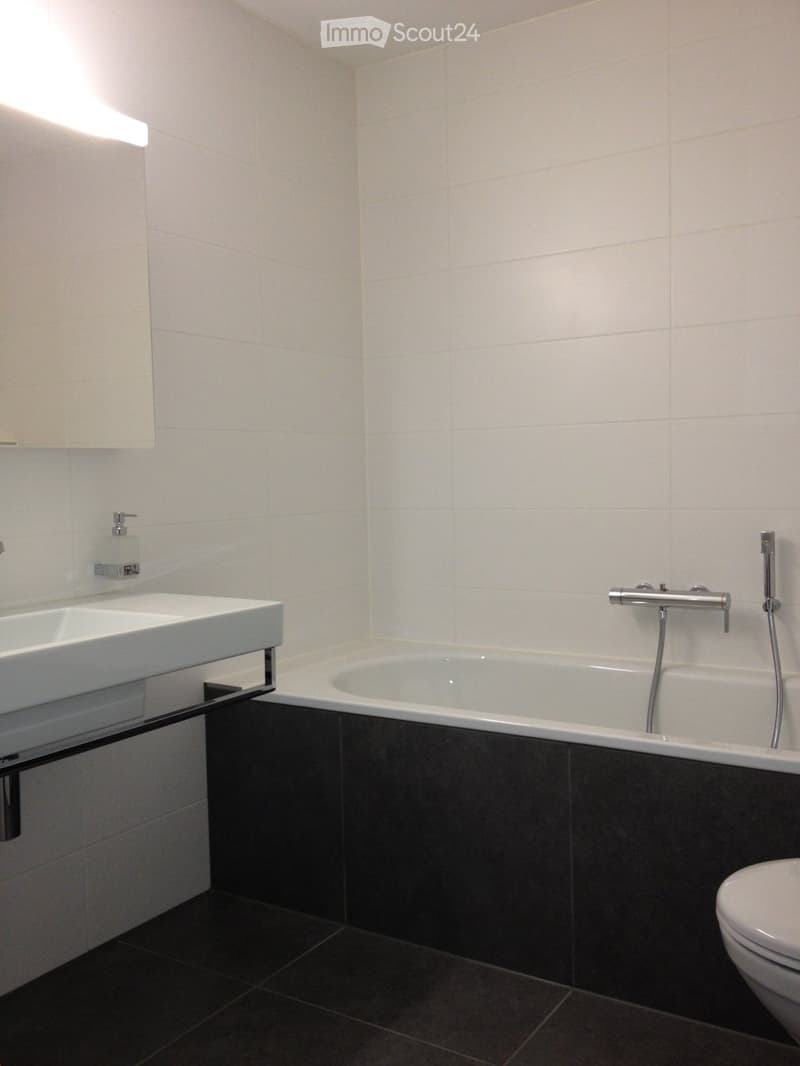 Modern bathroom with white tiles, a white sink, a bathtub, and a toilet. The walls are tiled in white, and the floor is dark grey.
