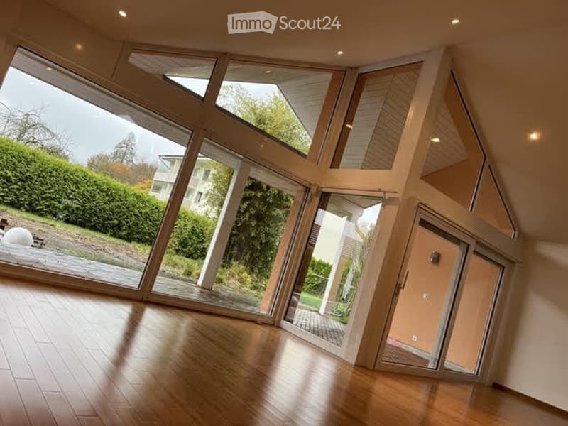 Bright, open-plan room with large glass walls and wooden flooring, featuring a modern, angular design and ample natural light.