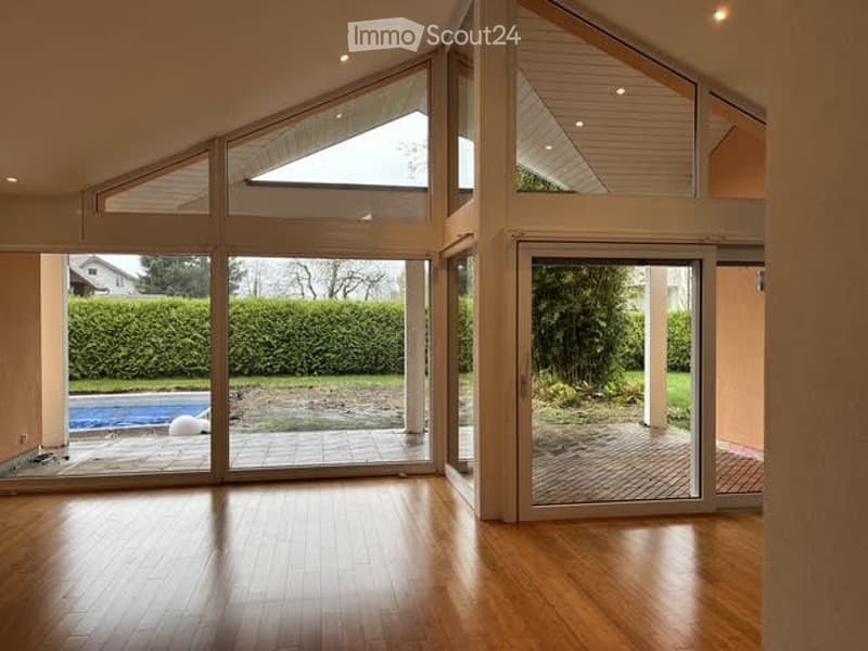 Bright, open-plan room with wooden flooring, large glass sliding doors leading to a patio and pool area, visible garden and swimming pool outside.