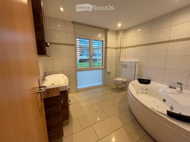 Modern bathroom with white tiles, a large bathtub, a toilet, a sink with a wooden cabinet, and a window with blinds.