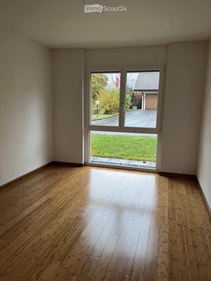 Empty room with wooden flooring and large sliding glass door leading to a green lawn and driveway.