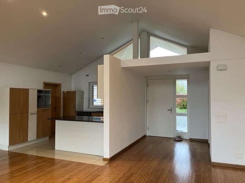 Open-plan living and kitchen area with wooden flooring, white walls, and modern kitchen cabinets. Features a central island with a countertop and an oven, along with a large door leading outside.