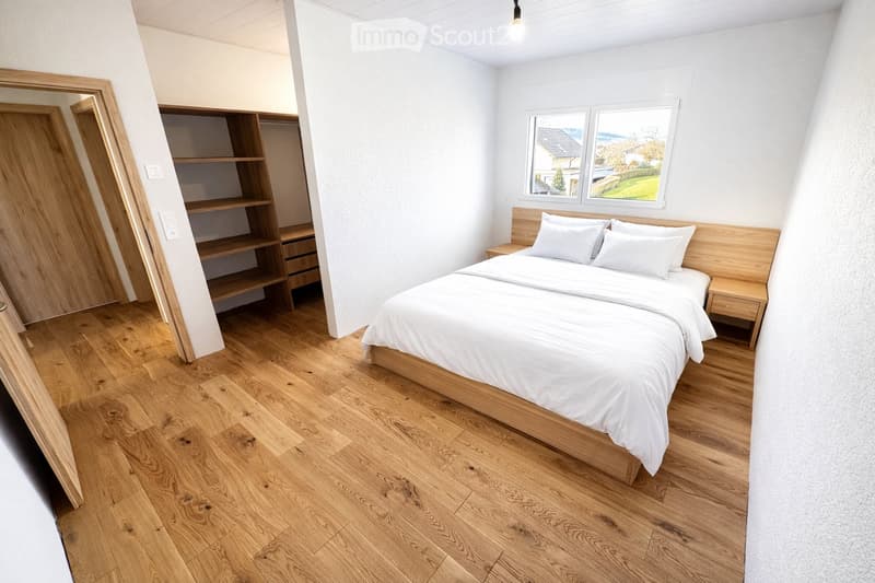 Modern bedroom with wooden flooring, a large bed with white bedding, a nightstand, a wardrobe with shelves, and a large mirror on the wall.