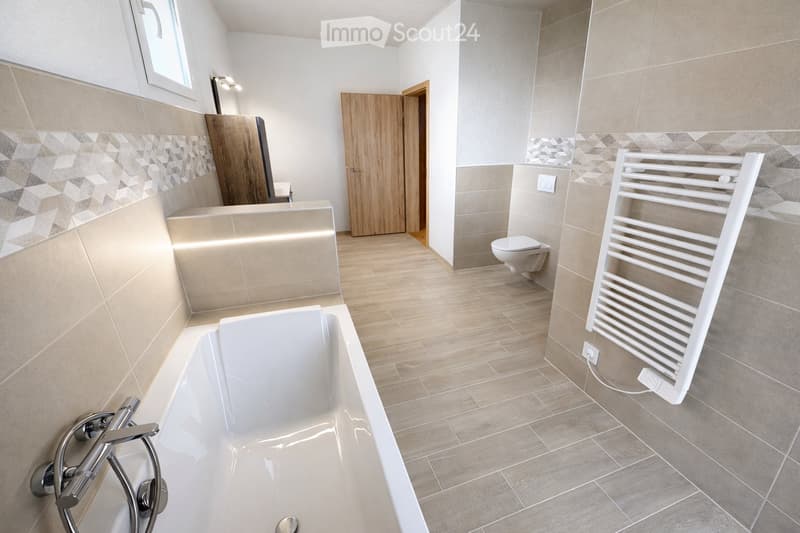 Modern bathroom with a white bathtub, toilet, radiator, and wooden door. Light-colored tiles and wooden flooring.