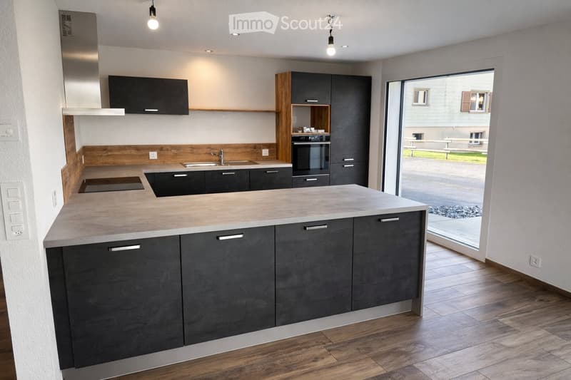 Modern kitchen with grey cabinets, a central island, and a sink. The space features wooden accents and a large window offering a view of the outdoor area.