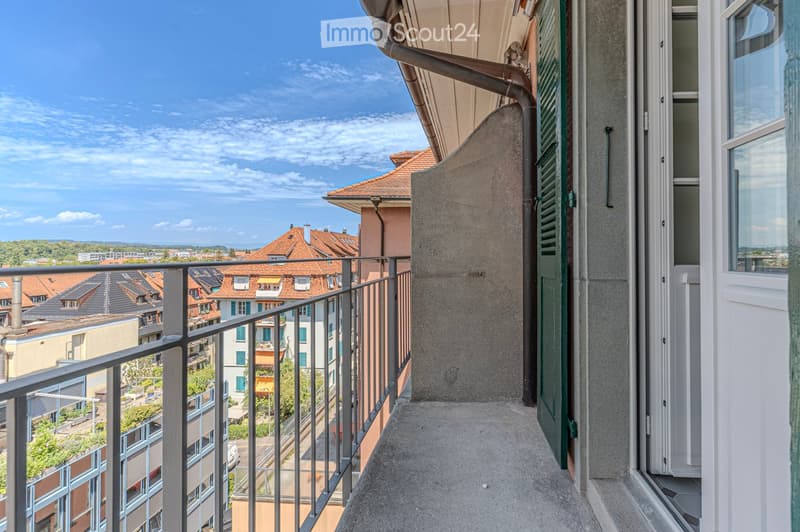 Balcony with metal railing, overlooking a cityscape with red-tiled roofs and greenery.