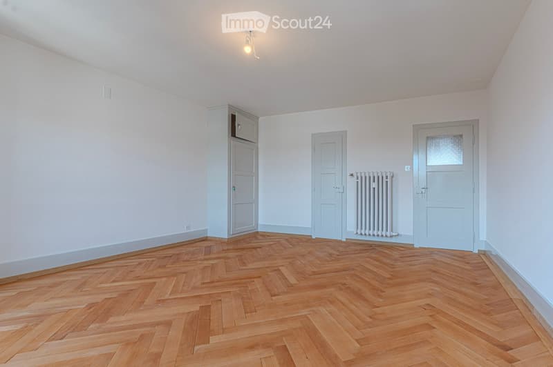 Empty room with wooden herringbone flooring, white walls, and three doors, including a closet with a small cabinet on top.