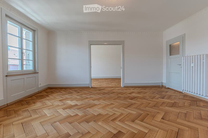 Empty room with wooden herringbone flooring, white walls, large window, and a door leading to another space.