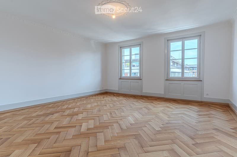 Empty room with wooden herringbone flooring, white walls, and two windows with radiators beneath them.