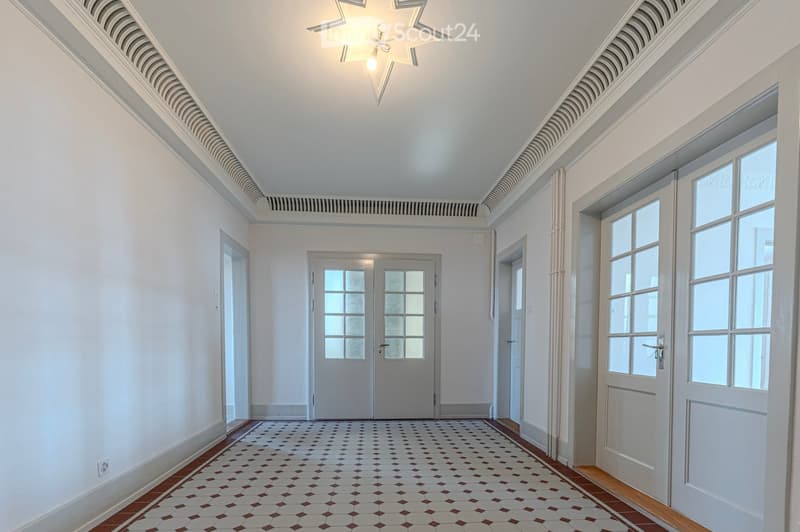 Bright, spacious hallway with white walls, decorative ceiling moldings, and a patterned tile floor. Features multiple doors and large windows allowing natural light to fill the space.
