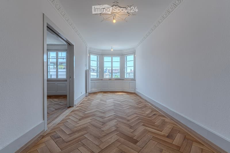 Empty hallway with herringbone wooden flooring, white walls, and ornate ceiling light fixture. Features a doorway leading to another room and large windows at the end.