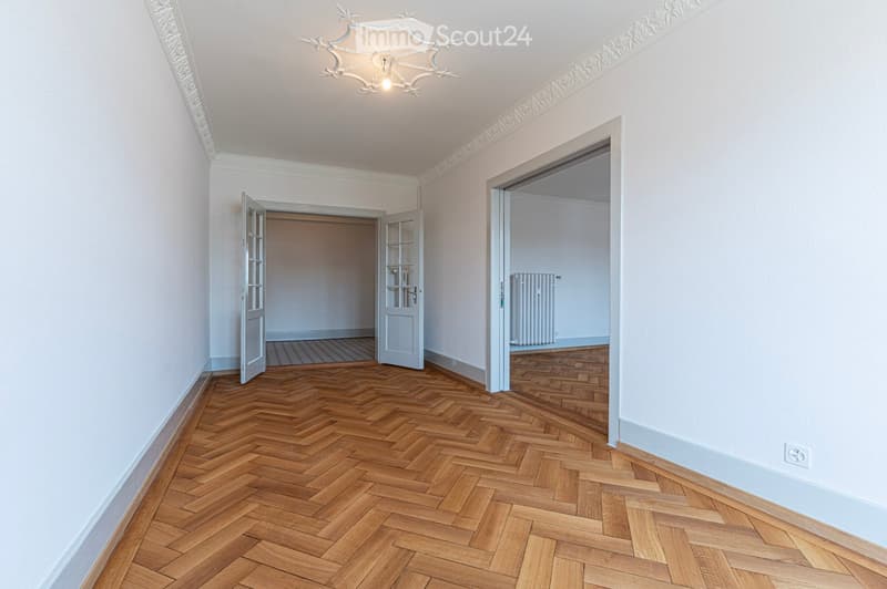 Empty room with wooden herringbone flooring, white walls, decorative ceiling molding, and two open doors leading to other rooms.