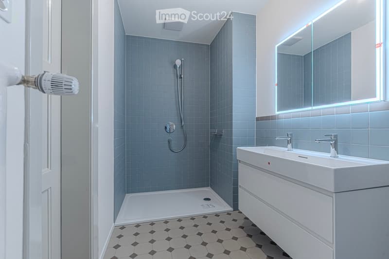 Modern bathroom with blue tiled shower area, white sink with dual faucets, large mirror with LED lighting, and black and white hexagonal floor tiles.