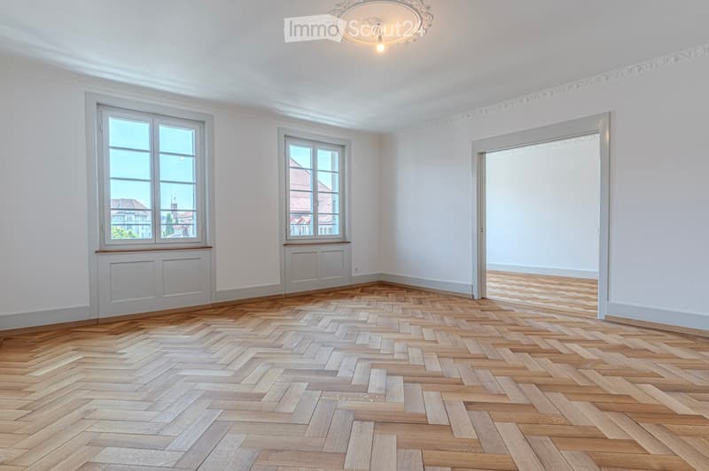 Empty room with wooden herringbone flooring, white walls, large windows, and a decorative ceiling light fixture.