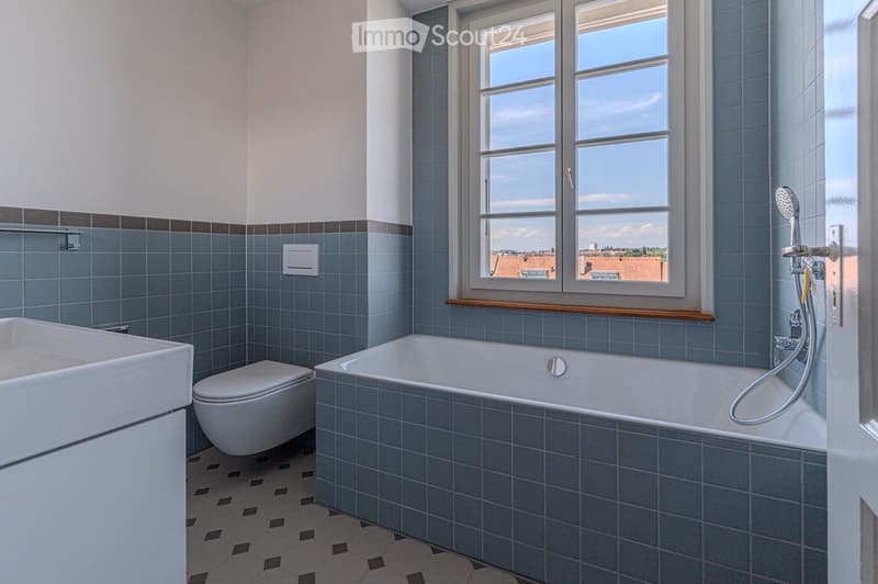 Modern bathroom with blue tiles, a bathtub, toilet, sink, and a large window offering a view of the city.