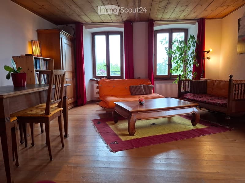 Cozy living room with wooden floors, large windows, a red curtain, a wooden coffee table, a sofa, a chair, a bookshelf, and potted plants.