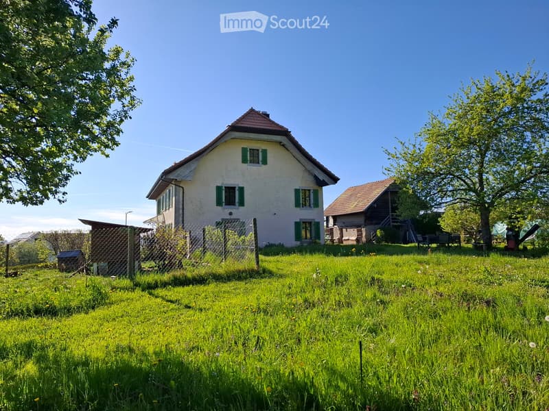 Two-story house with white walls, green shutters, and a red-tiled roof, surrounded by a grassy lawn and trees.