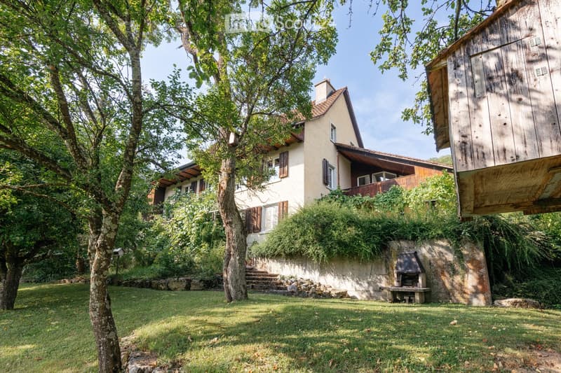 A two-story house with a beige facade, wooden shutters, and a stone chimney. The house is surrounded by lush greenery, including trees and shrubs, with a wooden outbuilding and a stone barbecue area nearby.