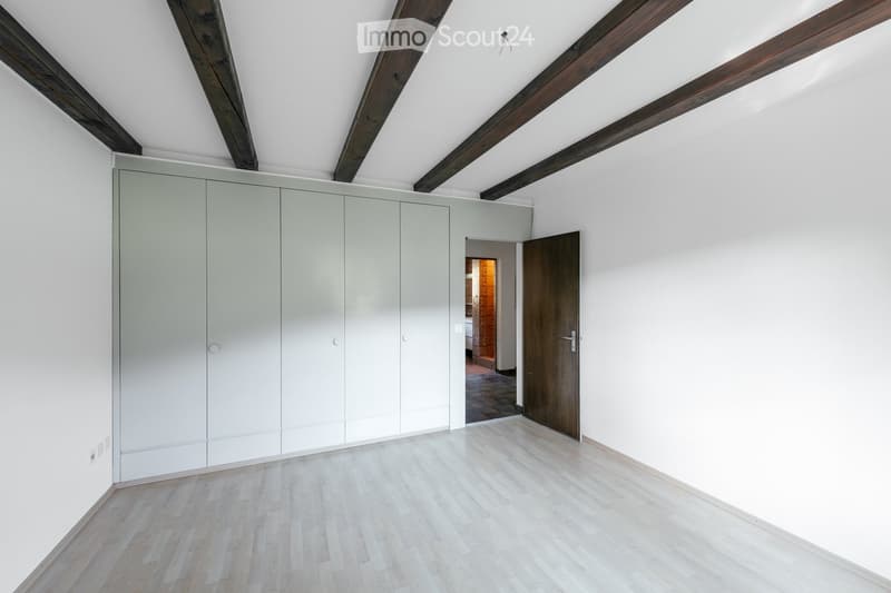 Empty room with light wooden flooring, white walls, exposed wooden beams on the ceiling, and a large built-in wardrobe unit. A doorway leads to another room with warm lighting.