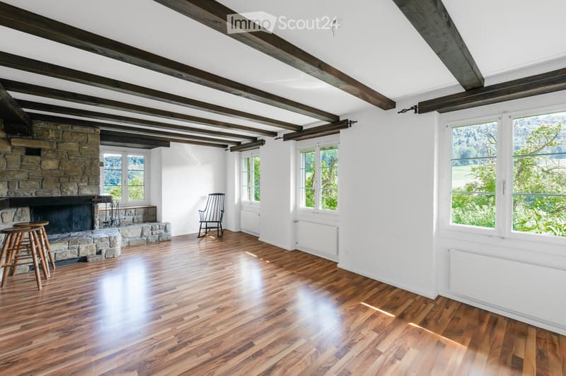 Bright living room with exposed wooden beams, hardwood flooring, large windows, and a stone fireplace.