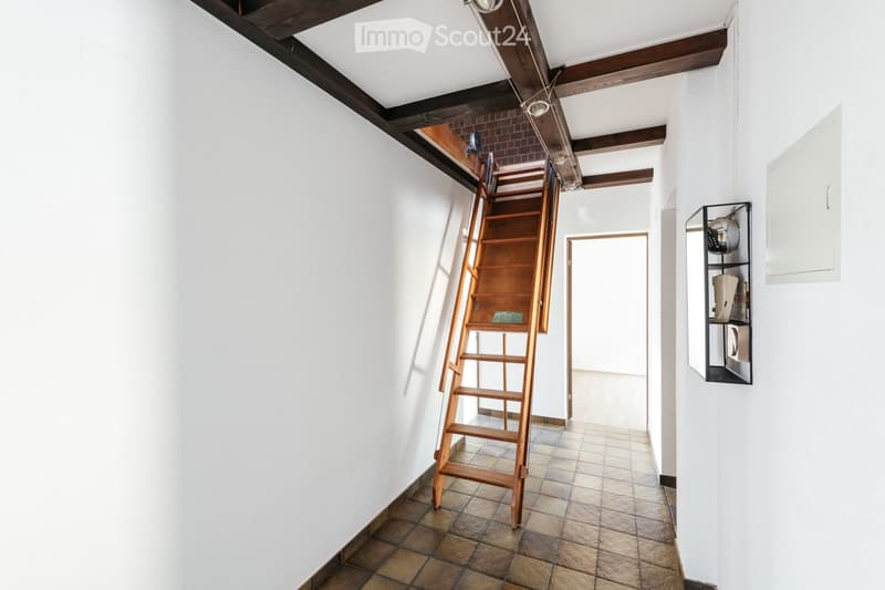 A narrow hallway with white walls, tiled floor, wooden ceiling beams, and a wooden ladder leading to an attic space. A small shelf with items is visible on the right side.