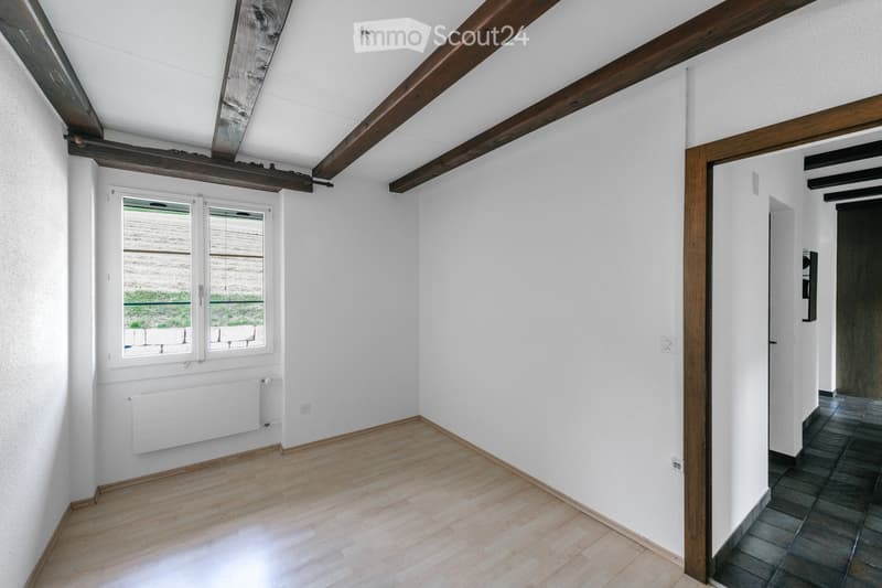 Empty room with wooden flooring, white walls, exposed wooden beams on the ceiling, and a window with a view of the outside.